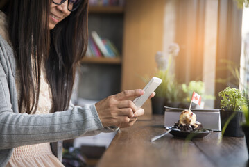 Beautiful woman wearing glasses take picture phone of cake (on wooden table) in cafe.Technology,food and drink concept.Bible and pot of tree on wooden table.