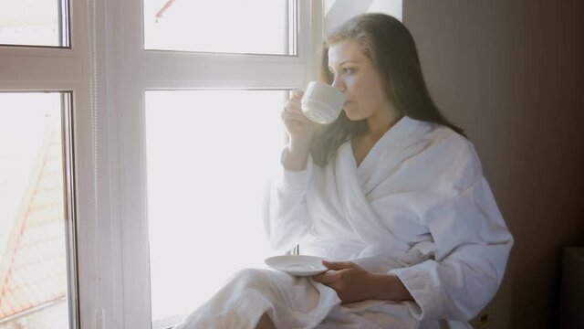 4k footage of young woman sitting on windowsill and drinking coffee