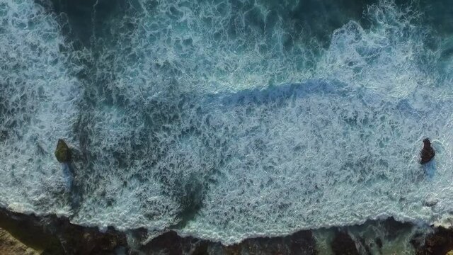 Tilt up aerial view from foamy waves crashing onto rocky shore to clear blue ocean water
