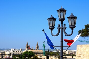 Maltese and EU flag on a lamppost with city buildings to the rear, Valletta, Malta.