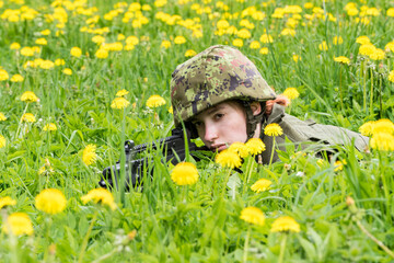 Portrait of armed woman with camouflage. Young female soldier observe with firearm. Child soldier with gun in war, green goutweed and yellow dandelions background.  Military, army people concept