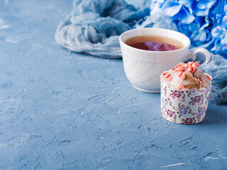 Obraz na płótnie Canvas Cup of tea on blue background with frosted cup cake, flowers and textile