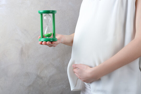 Young pregnant woman holding hourglass near belly on light wall background