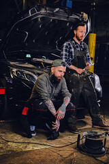 Two bearded tattooed mechanics near the car in a workshop.