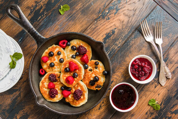 Homemade cottage cheese sweet pancakes with raspberry, black currant and blackberry in a cast-iron skillet and jam in white bowls on the wooden rustic table, top view.
