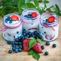 Three jars of yogurt with blueberries and strawberries, square. Berries of blueberries and strawberries are scattered on the table. Cooking, healthy food, food packaging
