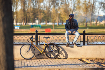 Vape in real life. Young man with gourgeous large beard and in sunglasses and in the cap having a rest and vaping an electronic cigarette near vintage fix bicycle after ride on the waterfront.