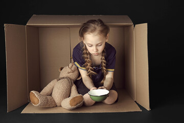 Poor little girl with empty bowl and toy bear sitting in cardboard box on black background