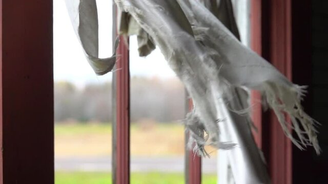 CLOSE UP, SLOW MOTION: Looking outside the shattered window in old ruined abandoned wooden hut. Spooky torn white curtain swaying in the wind. Spooky crumbling haunted house in American countryside