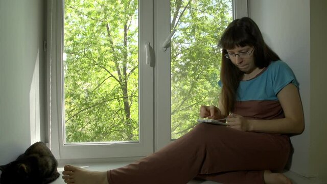 Young beautiful woman in glasses knits from blue yarn with knitting needles and uses a smartphone sitting on a windowsill at a bright spring day outside a tree with green leaves.