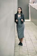 portrait of fashionable girl in long gray dress. Face brunette in black leather jackets with ruby lips on a background of stone