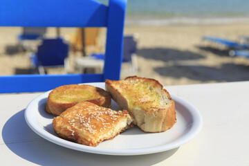 Bread with olive oil and feta cheese appetizer with honey and sesame seeds, Traditional greek dish on the white wooden table background served by the sea, Defocused chair and beach in the background