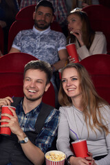 Smiling handsome boyfriend and beautiful girlfriend watching funny comedy movie at cinema together with friends, who sitting back. Positive friends holding red cup with cola and eating tasty popcorn. 