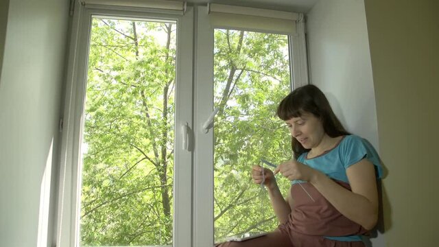 Young beautiful happy woman knits from blue yarn with knitting needles and having video chat , sitting on windowsill at home on bright spring day outside window trees with green leaves.
