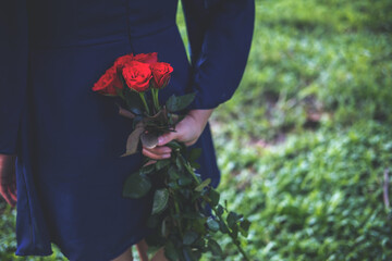 An Asian woman holding and hiding red roses bouquet on her back with green nature background