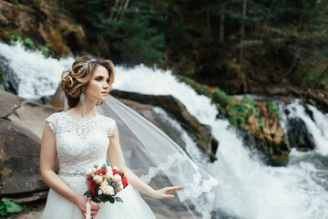 Wind blows bride's veil away while she stands before a waterfall