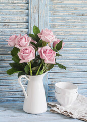 Pink roses in a white enameled pitcher and ceramic white bowls on blue wooden rustic background. Kitchen still life in vintage style