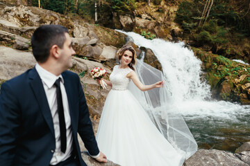 Bride in a magnificent dress stands behind her groom on the stones