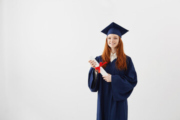Happy ginger graduate girl in cap and mantle smiling holding diploma. Copy space.