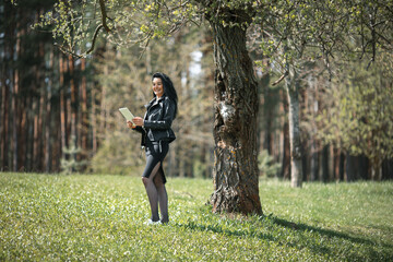 Gadget woman. Portrait of young pretty white girl with black hair and in black clothes surfing on the internet in white tablet near the old tree.