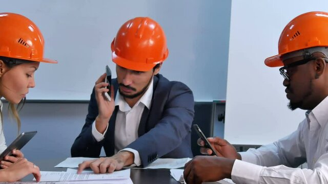 Tree colleagues in helmets discuss constraction plan, using digital tablet and smartphones
