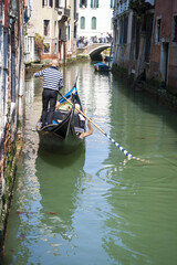 Fototapeta na wymiar Scenic view of a small canal in Venice, Italy, with gondolier in traditional striped shirt maneuvering gondola along green waters