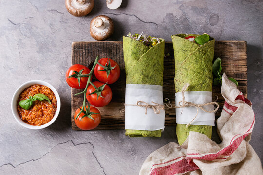 Green spinach matcha tortillas wrapped in paper with ingredients above. Sweet corn, avocado, green paprika, sprouts, mushrooms served on wood board over gray texture background and textile. Flat lay