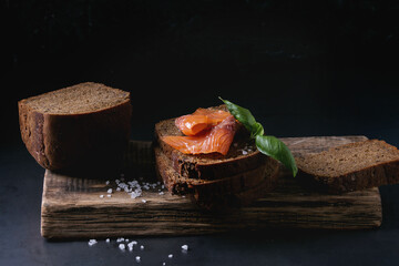 Stack of sliced homemade rye bread with smoked salmon, sea salt and fresh basil on wooden chopping board over dark black background.