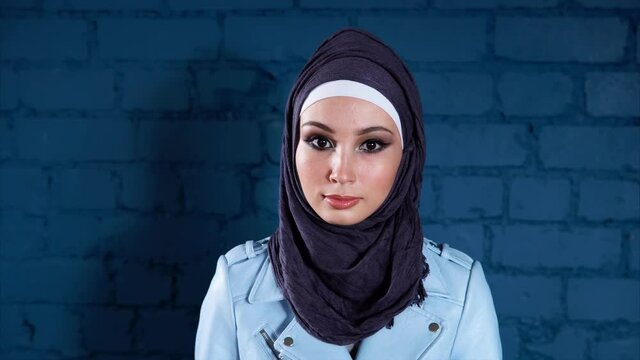 Young cheerful Muslim woman wearing black hijab is against blue brick wall background. Studio portrait