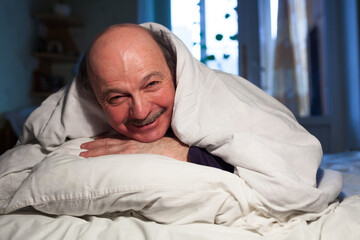 An elderly bald man with a mustache lies in bed, hugging a pillow. He has a problem with sleep.