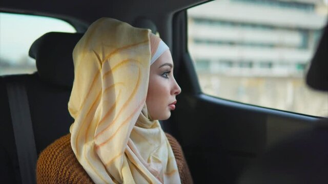 Young Muslim woman traveling on backseat of a taxi. She enjoying city view through the car window and laughing at the end