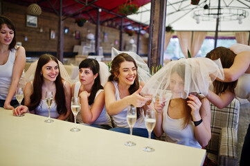 Group of cheerful girls at white shirts sitting at table and drink champagne on hen party.