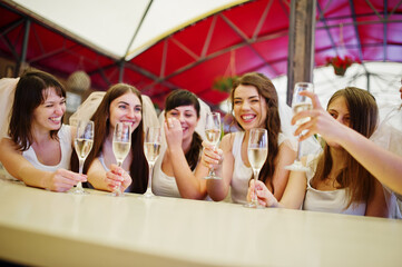 Group of cheerful girls at white shirts sitting at table and drink champagne on hen party.