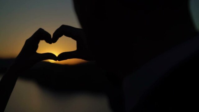 Young couple makes heart from hands on sunset near sea