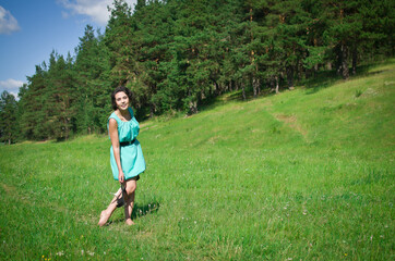 Young adult girl in shorts holding her summer shoes in her hand as she stands against rural green grass field and blue sky with clouds , close up view. full length