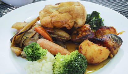 Traditional English Sunday lunch food of Yorkshire pudding, slices of meat, roast potatoes, carrots, broccoli, cauliflower, spinach, served on a white plate.