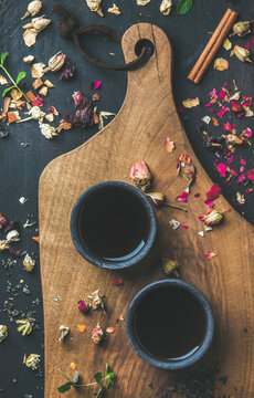 Chinese black tea in black stoneware cups on serving wooden board over black wooden background with herbs, flower buds, tea leaves spilt around, top view, vertical composition