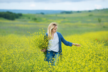 Genuine cute lady woman in meadow of yellow flowers sniffing flower bouquet. Attractive beautiful young girl enjoying the warm summer sun in a wide green and yellow meadow. Stylish dressed