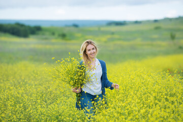 Fototapeta premium Genuine cute lady woman in meadow of yellow flowers sniffing flower bouquet. Attractive beautiful young girl enjoying the warm summer sun in a wide green and yellow meadow. Stylish dressed