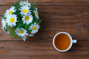 A bunch of chamomiles and a cup of tea on a wooden table, top view with a copy space.