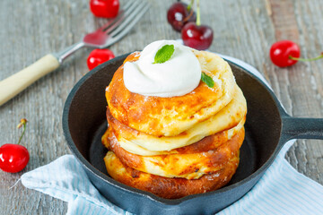Homemade cottage cheese pancakes with sour cream and cherries in a cast-iron frying pan. Russian dessert from cottage cheese.