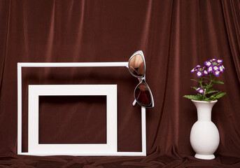 Two empty picture frames, glasses and vase with flowers on background of brown cloth.