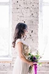 Fototapeta premium beautiful sexy Oriental girl brunette bride in white wedding dress with Golden sequins, a bouquet on a brick background in Studio near the window profile