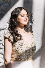 Fototapeta premium beautiful sexy Oriental girl in a white dress with a Golden glitter makeup long wavy hair standing on the hard sun with closed eyes smile close-up, portrait