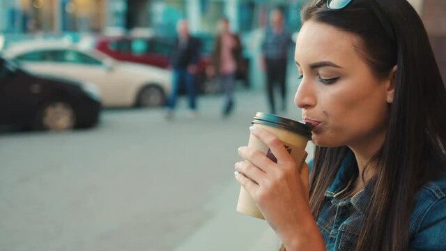 Attractive woman drinking coffee to go on the city street. Portrait shot. Woman sitting on the steps, drinking coffee and looking at the camera. Close up