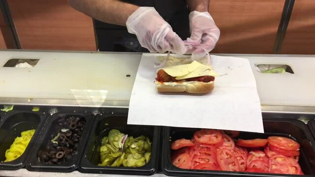Hands make small sub sandwich with cheese and light vegetables under bright lighting.  