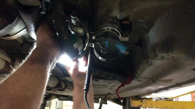 Mechanic holds bright light under car as camera moves slowly about capturing him repairing the fuel pump.