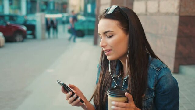 Beautiful young woman listening yo music on smart phone, singing and moving to the rythm while sitting on the steps outside on the street. Close up