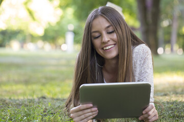 Fototapeta premium Young beautiful woman lying in the grass, looking at the tablet and enjoy nature