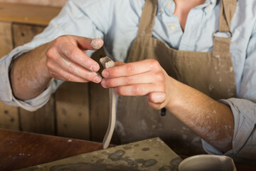 potter, stoneware, ceramics art concept - closeup on master hands working with some piece of clay, male works at a workshop near table, craftsman fingers forming the handle from raw fireclay
